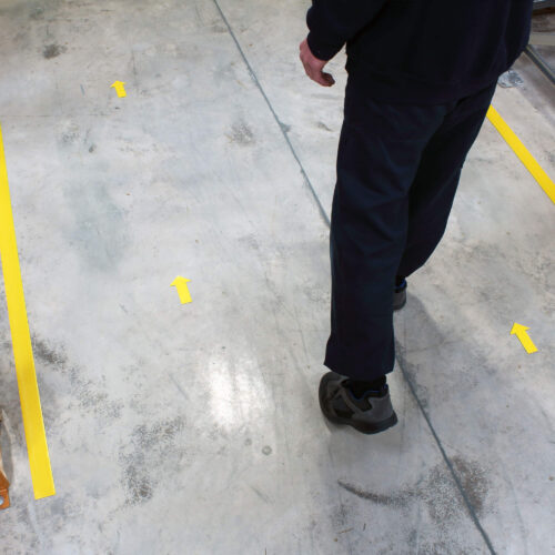 Worker walking on a concrete warehouse floor marked with yellow PermaStripe one-way aisle marking tape and directional arrows for safe pedestrian guidance.